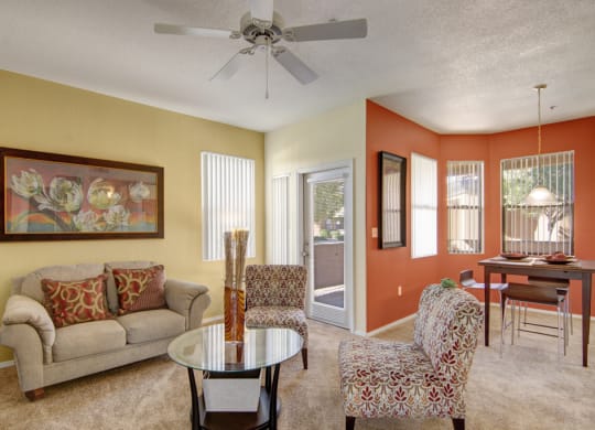 Bright living and dining area here at Arezzo Apartments with beige walls and an orange accent wall. Features a cozy sofa, patterned chairs, a glass coffee table, and modern decor.