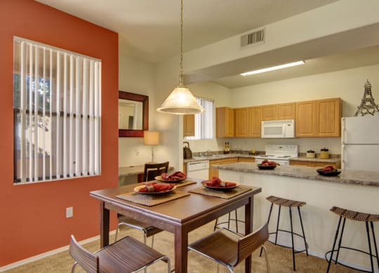 Warm kitchen-dining area here at Arezzo Apartments with a wooden table set for four. Beige cabinets, white appliances, and a bar with stools create a cozy, inviting vibe.