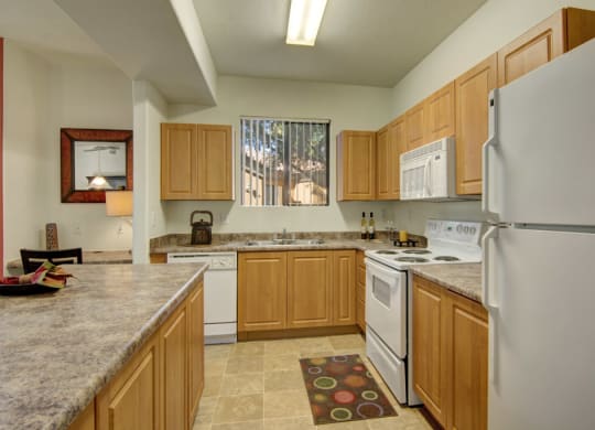 Bright kitchen here at Arezzo Apartments with wooden cabinets, white appliances, and granite countertops. A colorful rug lies on tiled floor; natural light from a window adds warmth.