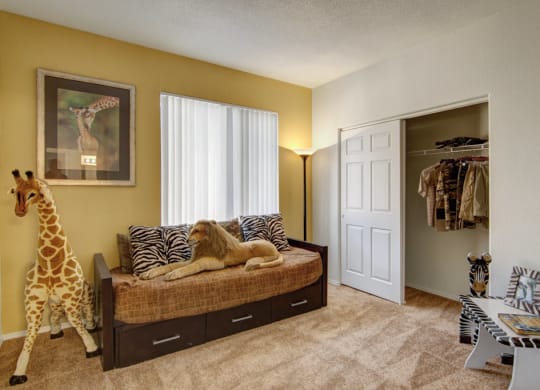 Cozy bedroom here at Arezzo Apartments with safari theme; features a giraffe statue, daybed with lion and zebra cushions. Warm tones, open closet, zebra-print side table.
