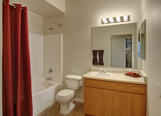 A tidy bathroom here at Arezzo Apartments with beige walls features a white bathtub with red curtains. There's a wooden cabinet with a white sink, a large mirror, and bright overhead lights.