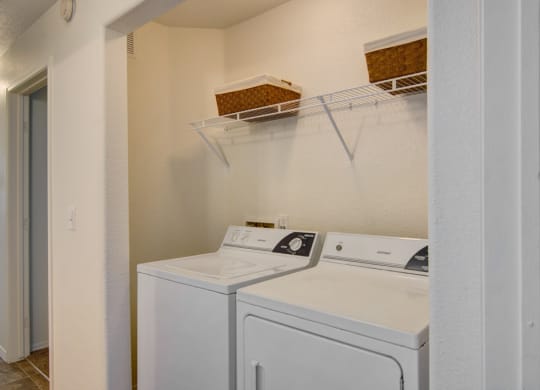 Laundry area here at Arezzo Apartments with a washer and dryer, topped with a metal shelf holding two brown baskets. The space is clean and organized, with a simple white interior.
