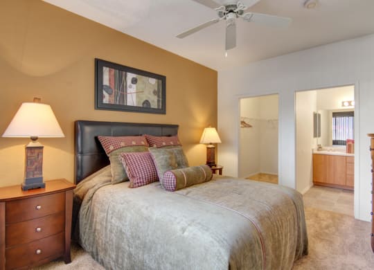 Cozy bedroom here at Arezzo Apartments with beige walls, a plush bed with patterned pillows, two wooden nightstands with lamps, a ceiling fan, and an en-suite bathroom.