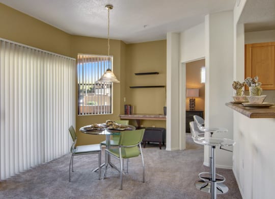 Modern dining area here at Arezzo Apartments with round table, green chairs, and pendant light. Large window with vertical blinds, bar with stools, and adjacent room visible. Cosy and inviting atmosphere.