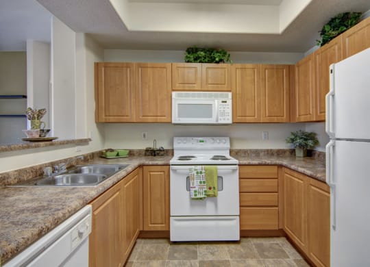 A cozy kitchen here at Arezzo Apartments with light wood cabinets, gray countertops, a white stove and microwave, and a fridge. Green plants add a fresh touch to the decor.