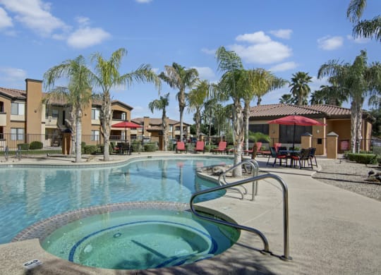 Resort-style pool area here at Arezzo Apartments with a jacuzzi in the foreground, surrounded by palm trees. Red umbrella and chairs sit on a sunny poolside patio.