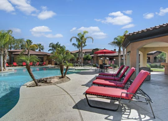 Sunny poolside scene here at Arezzo Apartments with red lounge chairs, palm trees, and clear blue sky. Relaxing atmosphere with patio area and umbrellas in a resort setting.