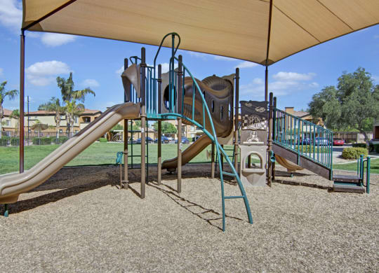Playground here at Arezzo Apartments with slides and climbing structures under a shade canopy. Surrounded by grass, trees, and residential buildings on a sunny day.