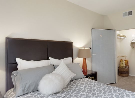 Bedroom interior here at Acclaim Apartments with a neatly made bed, decorative pillows, and a dark headboard. A modern lamp on a side table, and an open closet with baskets conveys a cozy, organized atmosphere.