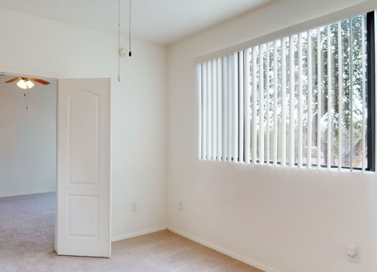 Empty room here at Acclaim Apartments with white walls and beige carpet, featuring a half-open door leading to another room with a ceiling fan. Vertical blinds cover a window on the right. Bright and minimalistic.