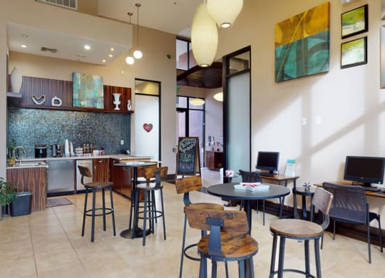 Modern lounge area here at Acclaim Apartments with wooden high tables, stools, and computers. Features a kitchenette with mosaic tiles, contemporary art, and large pendant lights.