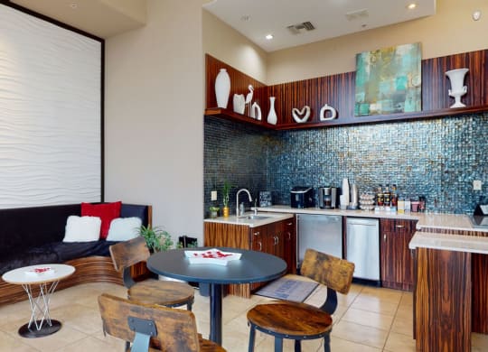 Modern kitchen lounge here at Acclaim Apartments with wood-accented cabinets, black and white decor, wavy textured wall, blue tile backsplash, and cozy seating with red pillows.