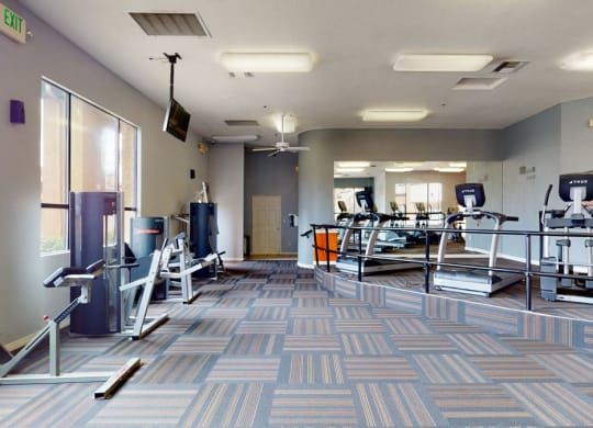 Modern gym interior here at Arezzo Apartments with exercise machines, including ellipticals and weight stations, on patterned carpet. Bright, spacious, with large mirrors and windows.