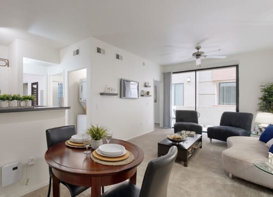 Modern living room here at Bolero Apartments with neutral tones, featuring a round dining table set for two, cozy gray seating, a kitchen with potted plants, and large windows.