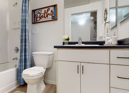 A clean, modern bathroom here at Bolero Apartments with a white bathtub and blue plaid shower curtain. The vanity has a black countertop, a sink, and soap dispenser. A wooden sign reads "Hot Baths."