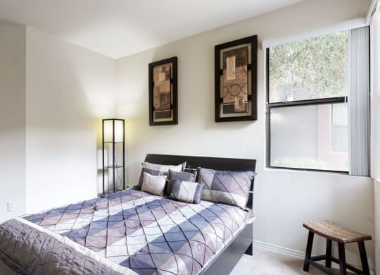 A cozy bedroom here at Bolero Apartments with a geometric-patterned bedspread and cushions. Two abstract paintings hang above. A window with a view and soft lighting enhance the calm ambiance.