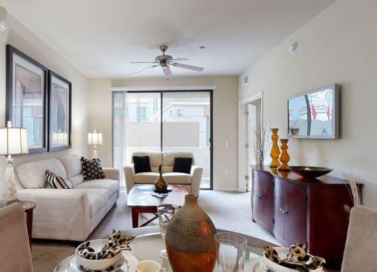 A modern living room here at Bolero Apartments with neutral tones, a white sofa, patterned cushions, and a wooden coffee table. Large windows bring in light, enhancing the airy ambiance. A ceiling fan and framed artwork decorate the walls, while assorted vases and decor add warmth.