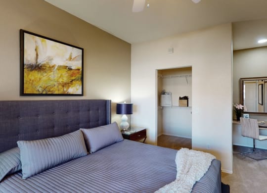 Cozy bedroom here at Dobson Towne Centre Apartments with a gray upholstered bed, striped bedding, and abstract art. A lamp and nightstand are beside the bed. A closet and vanity area are visible.