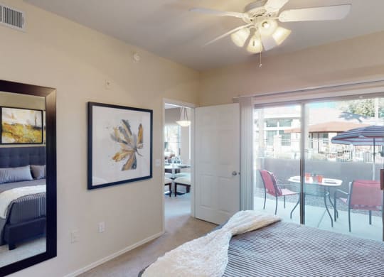 A cozy bedroom here at Dobson Towne Centre Apartments with a striped bedspread, mirror reflecting abstract wall art, and a ceiling fan. Sliding doors open to a patio with red chairs and an umbrella.