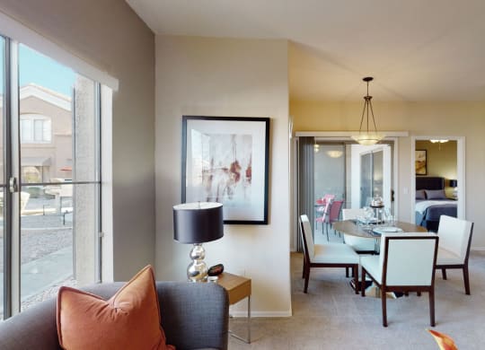 Modern living room here at Dobson Towne Centre Apartments with a grey couch and orange pillow, a framed abstract painting, and a lamp. Dining area visible with a round table, chairs, and soft lighting.