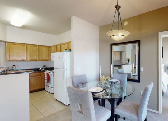 Dining area here at Dobson Towne Centre Apartments with glass table, four gray chairs, and a large wall mirror. Adjacent kitchen has light wood cabinets and white appliances. Bright, cozy atmosphere.