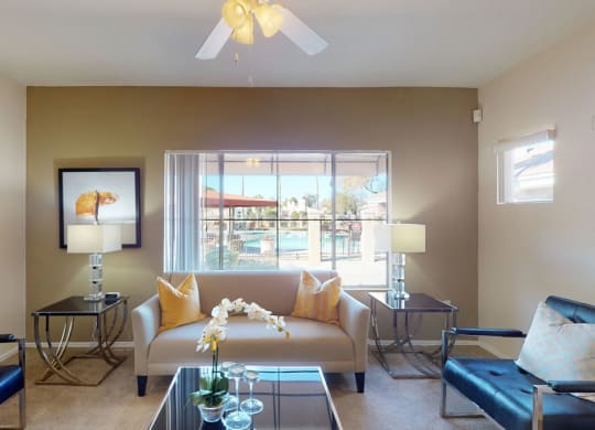 Stylish living room here at Dobson Towne Centre Apartments with a beige sofa, yellow pillows, and modern chairs facing a glass coffee table. Large window reveals a sunny poolside view. Neutral tones.