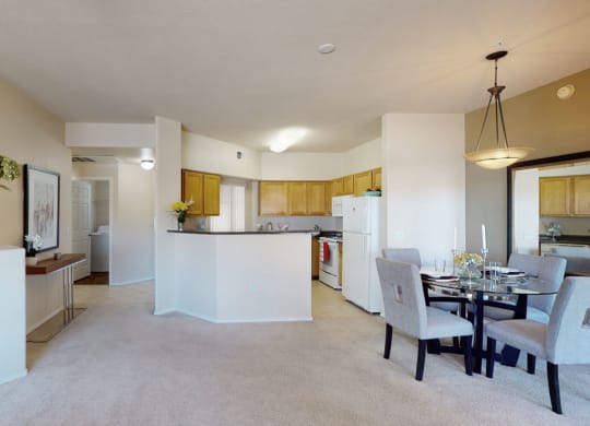 Modern living room here at Dobson Towne Centre Apartments with beige walls and carpet, open to a kitchen with wooden cabinets. A glass dining table with gray chairs sits under a pendant light.