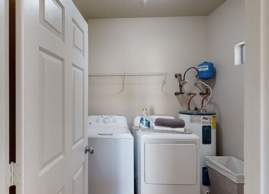 A small laundry room here at Dobson Towne Centre Apartments with a washing machine and dryer. A shelf above holds a detergent bottle and folded towels. A water heater is to the right.