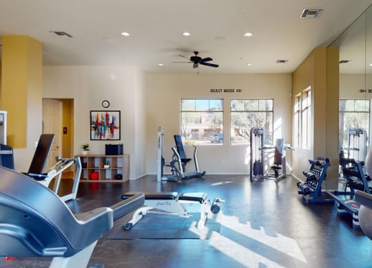 Bright gym here at Dobson Towne Centre Apartments with various equipment, including stationary bikes, dumbbells, yoga mats, and an exercise ball. "BEAST MODE ON" sign adds motivation. Calm and inviting atmosphere.