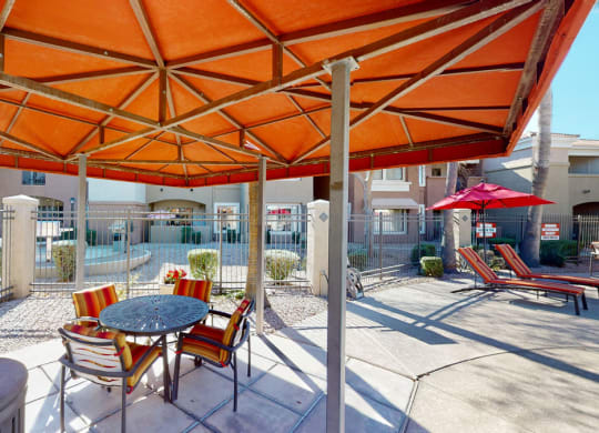 Outdoor patio here at Dobson Towne Centre Apartments under an orange canopy with a round table and striped chairs. Nearby, pool loungers with red umbrellas. Sunny and inviting ambiance.