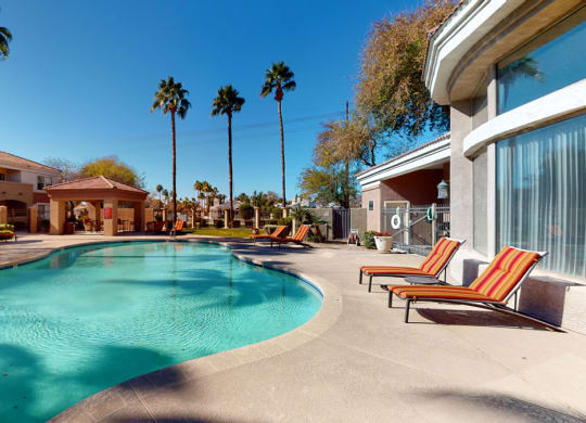 Sunny outdoor pool area here at Dobson Towne Centre Apartments with clear blue water, surrounded by palm trees. Red-striped lounge chairs line the pool deck, creating a relaxing atmosphere.