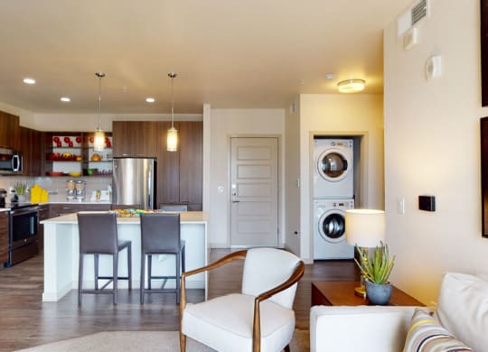 Modern open-plan kitchen and living area here at Escape Apartments with wood cabinets, stainless steel appliances, island with bar stools, cozy seating, and colorful wall art.