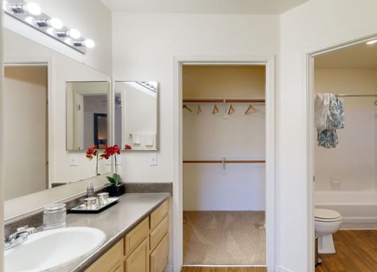Bright bathroom here at Mirador Apartments with a vanity, mirror, and vibrant flowers on the counter. Open closet with hangers and an adjacent shower area with wood flooring.