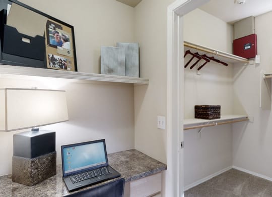 Cozy nook with a laptop on a granite desk here at Natura Villas Apartments, lit by a modern lamp. Nearby, an open closet shows hangers and a wicker basket on shelves.