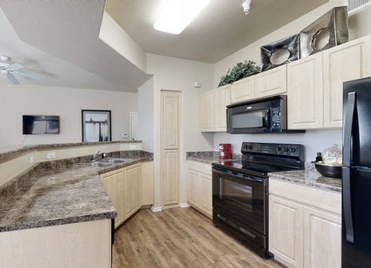 Modern kitchen here at Natura Villas Apartments with light wood cabinets, dark countertops, and black appliances including a stove and fridge. Open layout with a cozy, inviting ambiance.