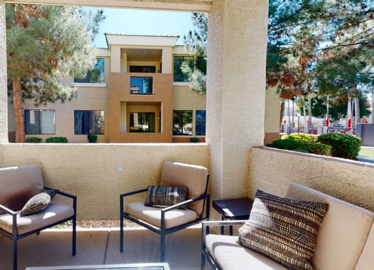 A cozy patio here at Natura Villas Apartments features beige armchairs with cushions, set against the backdrop of a stucco wall. Beyond, a modern apartment building and trees create a tranquil atmosphere.
