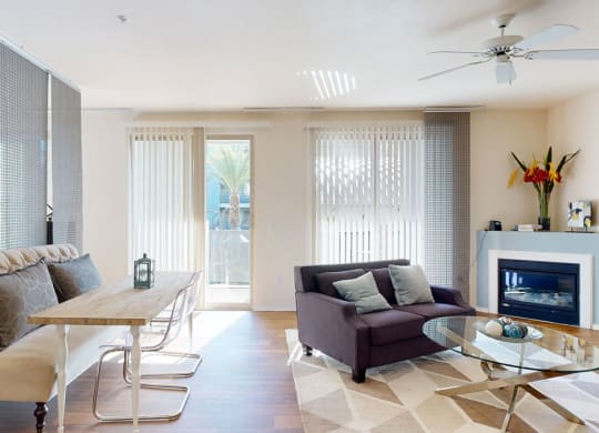 Modern living room here at Rio Paradiso Apartments with natural light, featuring a gray sofa, glass coffee table, dining bench, and decorative fireplace. Minimalist and cozy atmosphere.
