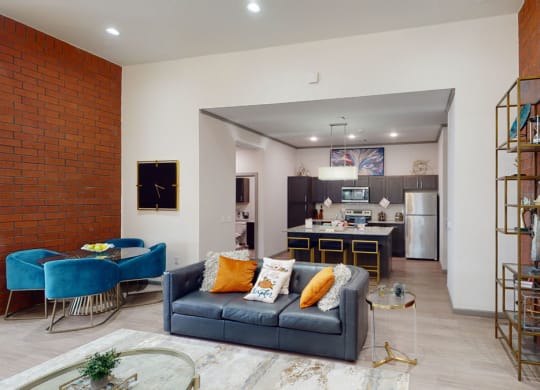 Contemporary resident lounge with a blue couch, orange and white pillows, and a glass table. A modern kitchen with dark cabinets is visible in the background.