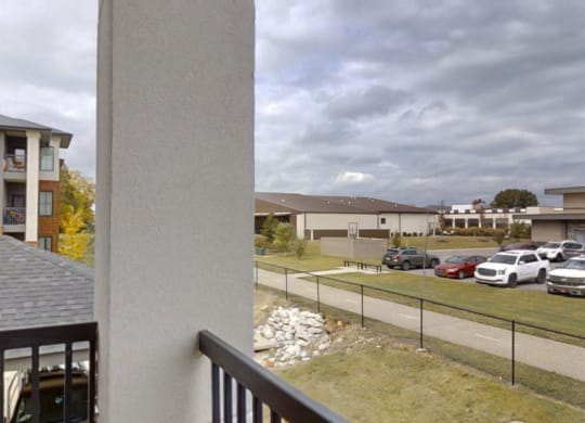 View from a balcony here at 49 West Apartments overlooking a parking lot with several cars, a fenced pathway, and nearby modern buildings under a cloudy sky, conveying a calm, suburban atmosphere.