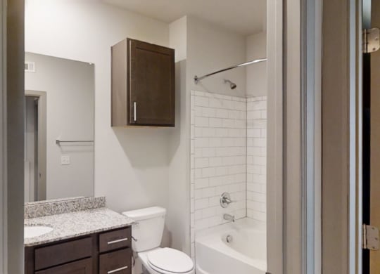 Modern bathroom here at 49 West Apartments with white tile bathtub and shower, dark wood cabinets, granite countertop, large wall mirror, and neutral walls. Clean and minimalistic.