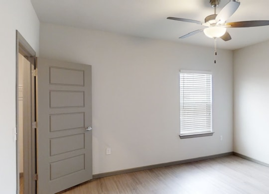 Empty bedroom here at 49 West Apartments with light gray walls and wooden floor, featuring an open door on the left, a closed window with blinds, and a ceiling fan with light.