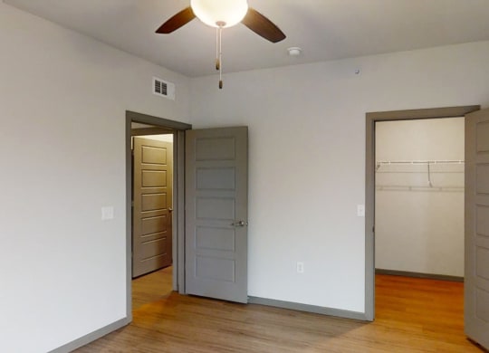 Empty bedroom here at 49 West Apartments with light gray walls and wooden floor. Two open doors reveal a closet and another room. A ceiling fan with a light fixture hangs in the center.