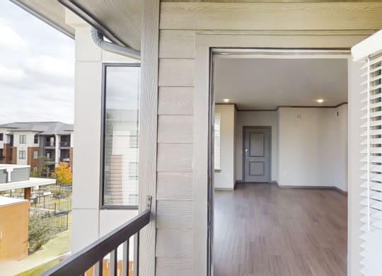View from a balcony here at 49 West Apartments into an empty apartment with wood flooring, neutral walls, and a closed door. Nearby buildings and trees are visible outside.