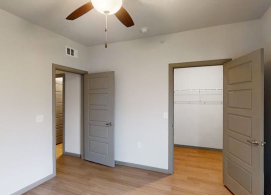 A minimal bedroom here at 49 West Apartments featuring light gray walls, a wooden floor, two open doors, and a ceiling fan with lights. Bright, serene, and spacious ambiance.