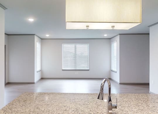 Spacious, modern room here at 49 West Apartments with light gray walls and large windows featuring white blinds. Granite countertop with sleek faucet in foreground, pendant lighting above.