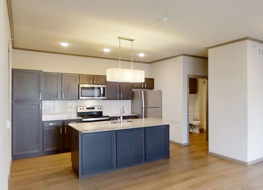 Modern kitchen here at 49 West Apartments with dark cabinetry, stainless steel appliances, and a granite island under pendant lighting; open layout with wood flooring.