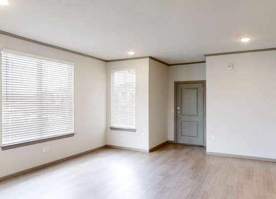 A modern, empty living room here at 49 West Apartments with light wood flooring, white walls, and two large windows with blinds. A closed gray door is on the right. Bright and minimalist.