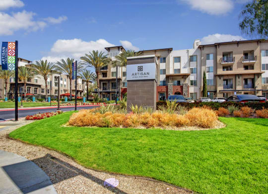 Modern apartment complex with lush green lawn in foreground. Sign reads “Artisan at Main Street Metro.” Palm trees and colorful banners line the street, under a blue sky.
