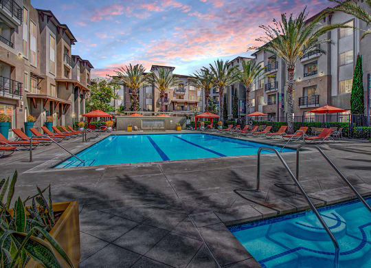 Modern Artisan at Main Street Metro apartment complex at sunset with a central pool, surrounded by palm trees and lounge chairs. The sky is vibrant with pink and blue hues, creating a relaxing ambiance.