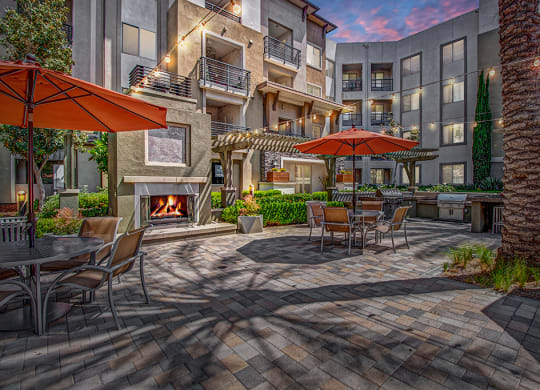 Cozy outdoor patio here at Artisan at Main Street Metro Apartments with tables, orange umbrellas, and string lights. A lit fireplace adds warmth. Surrounded by modern apartments at dusk.
